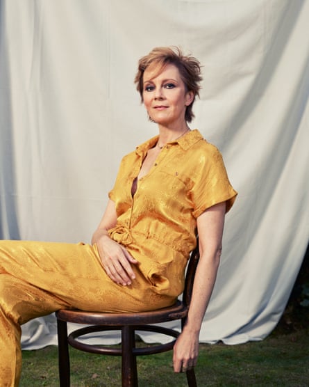 Rachael Stirling is wearing a yellow top and trousers, sitting on a wooden chair against a white curtain backdrop