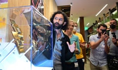 A man gestures near a transparent box containing computer components as people look on and take photos.