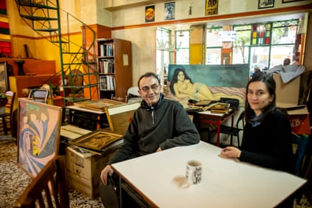 Christos Vanas and his daugher Areti sit at a table with a mug of coffee in front of them. There are paintings propped in the background behind them, including one of a reclining nude, and a metal spiral staircase. The walls are painted cream and yellow, with a red stripe, and there is stained glass in the windows.