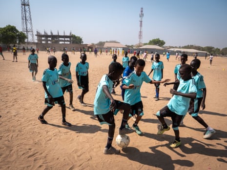 Young boys in a light blue team shirt, playing football on a sand pitch