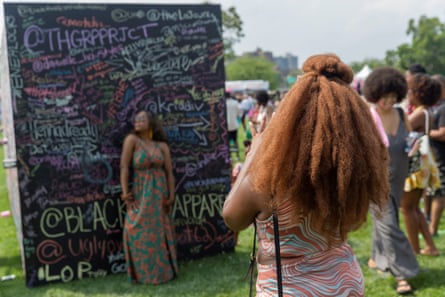 A woman poses against a chalkboard covered in slogans and hashtags. A woman with long natural hair is taking the photo, and in the background women with a variety of natural styles are milling around