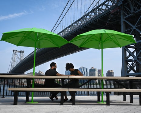 A couple sit at a table with green parasols under the Williamsburg Bridge looking over at Brooklyn