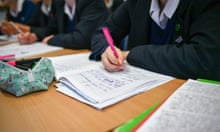 A student writes with a pink pen in an exercise book at a school desk in a classroom
