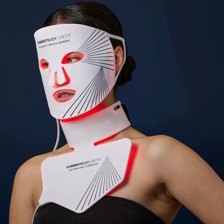 A woman with brown hair wears the white and black striped CurrentBody Skin LED Face & Neck Kit as it glows red, set to a dark blue background