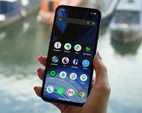 A hand holds a Fairphone 6+ showing its home screen, with boats blurred in the background
