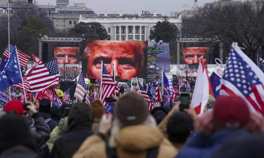 Apoiadores de Trump agitando bandeiras americanas participam do comício em 6 de janeiro em Washington DC. Três telas gigantes projetam um grande close do rosto de Donald Trump.