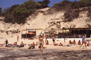 People gathered on a beach. A naked man is walking away from the camera in the centre of the shot
