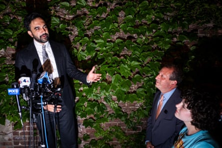 a man speaks into microphones while gesturing to another man standing nearby