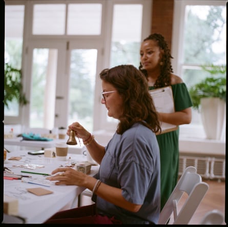 One lady is sitting at a table while another lady stands by her and observes
