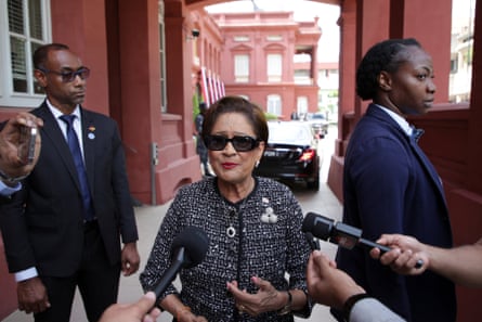 A short Asian woman in sunglasses flanked by bodyguards talking to the press