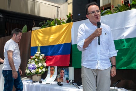 Jesús Abad Colorado speaks into a microphone as he stands next to a table with photographs of Mateo Pérez Rueda and a Colombian flag draped on a wall