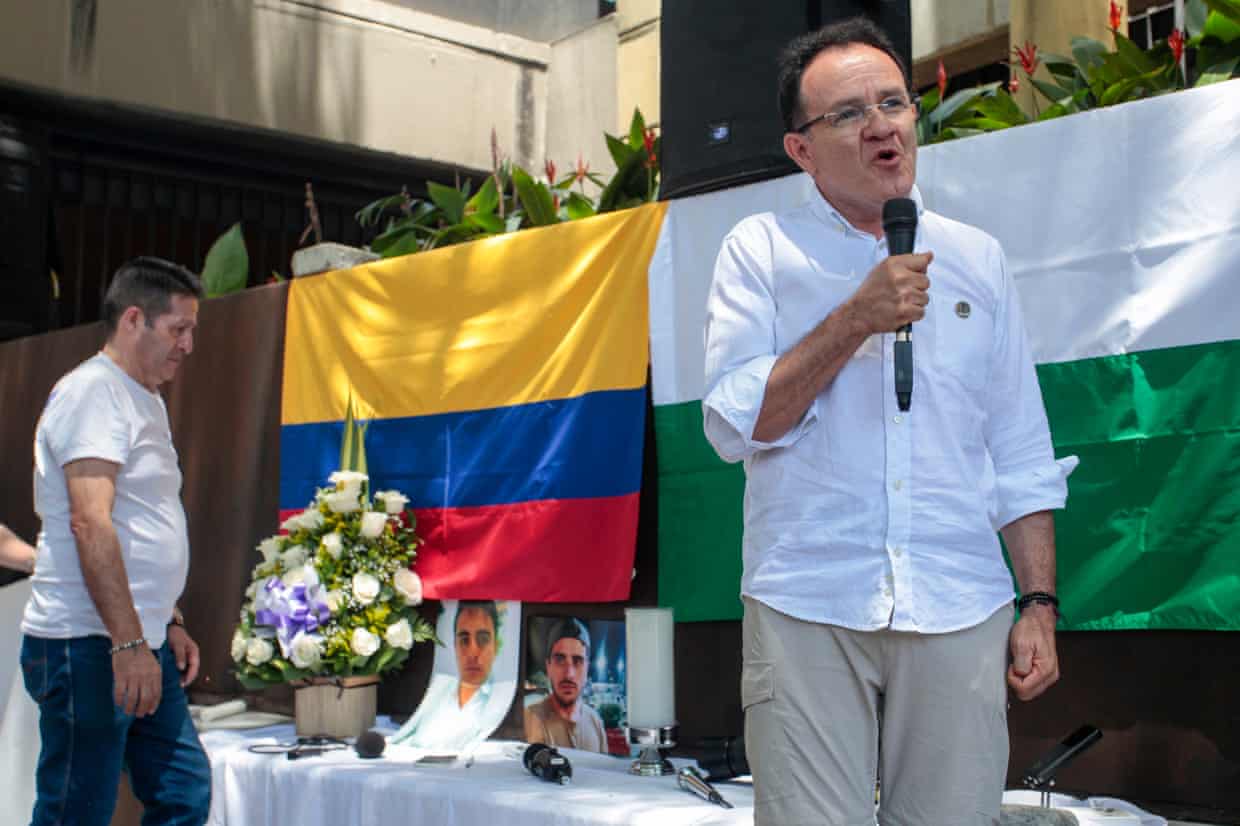 Jesús Abad Colorado speaks into a microphone as he stands next to a table with photographs of Mateo Pérez Rueda and a Colombian flag draped on a wall