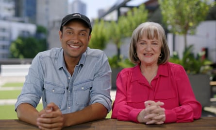 Comedians Matt Okine and Denise Scott sitting next to one another. Okine is wearing a faded blue shirt and a black cap while Scott is wearing a bright pink shirt.