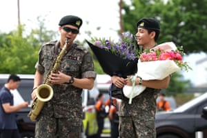 A man in military fatigues, sunglasses and a black beret plays the saxophone while another man similarly dressed stands beside him holding bunches of flowers
