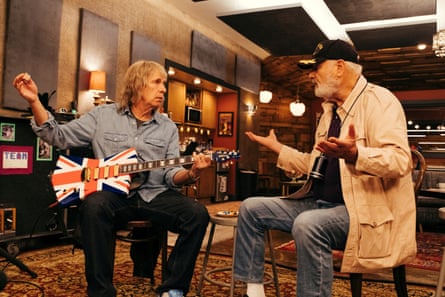 A man playing guitar talks to another man in a recording studio