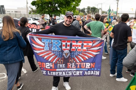 A man holds up a flag with the slogan: “Trump – 2024 THE RETURN. MAKE LIBERALS CRY AGAIN.”