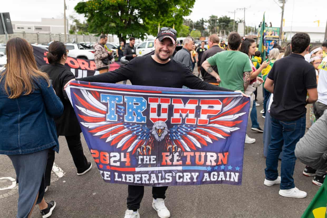 A man holds up a flag with the slogan: “Trump – 2024 THE RETURN. MAKE LIBERALS CRY AGAIN.”