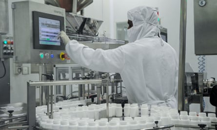Man in full biosecurity suit in a pharmaceutical plant fiddling with a screen while a production line of medicines operates around him.