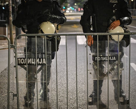 Riot police with helmets and shields stand behind barriers on a Belgrade street at night
