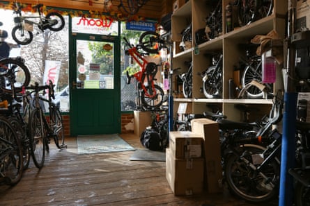 Interior view of the bike shop through to the front window, with folding bicycles in wooden shelving, and others hanging from the ceiling, and stacked to one side. There are e-bikes and children’s bikes.