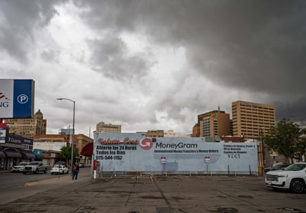 A wide shot depicts the side of a store advertising its international money transfer services, situated among other businesses, with a cityscape in the background