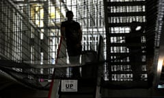 a general view of a prison: two men seen silhouetted as light streams through barred gates, walkways and metal stairways. A sign is seen reading 'this way up at mealtimes'.