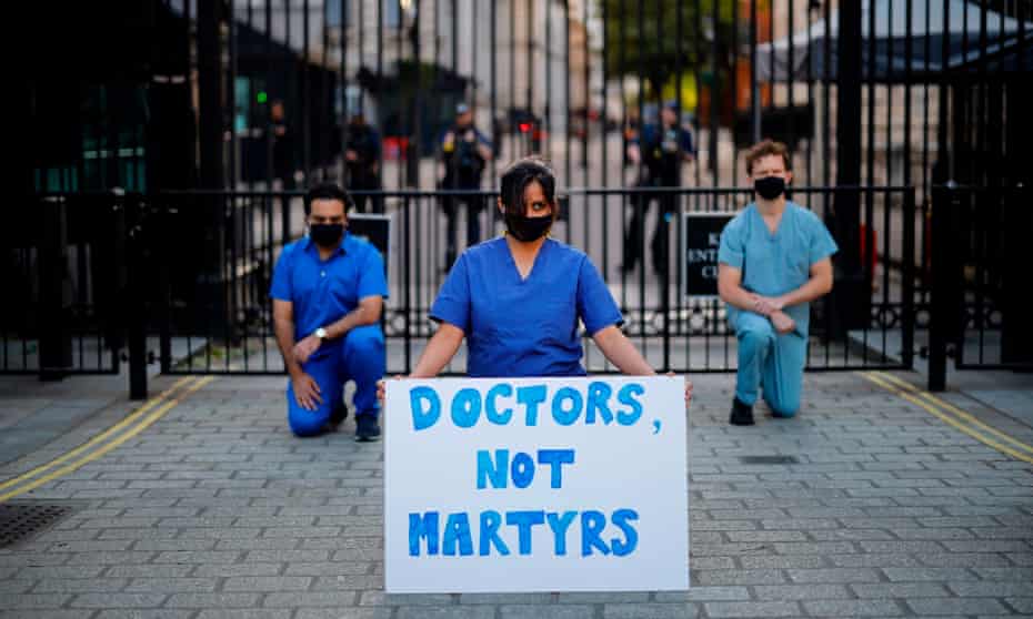 Three doctors in scrubs and masks kneeling outside the Downing Street gates with one holding a placard reading ‘Doctors Not Martyrs’