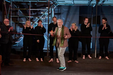 Ian McKellen stands holding scissors to cut the red ribbon as cast members applaud behind him.