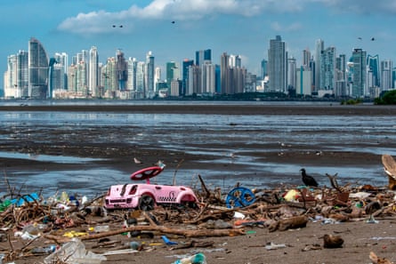 Cormorants pick for food among debris including a faded pink child’s pedal car washed up on a beach with other plastic waste and wooden flotsam. A skyline of tower blocks can be seen across mudflats 