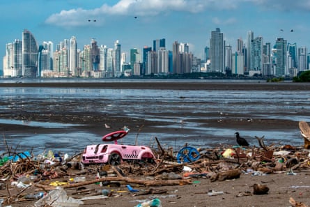 Cormorants pick for food among debris including a faded pink child’s pedal car washed up on a beach with other plastic waste and wooden flotsam. A skyline of tower blocks can be seen across mudflats