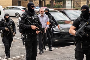 Heavily armed police in balaclavas escort a man in handcuffs across a car park