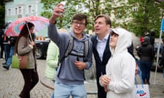 Maximilian Krah, in a shirt and suit jacket and holding a shopping bag printed with "AfD", poses with two young men as one holds up his phone for a selfie