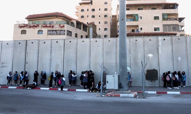 Palestinian schoolgirls wait for buses in the shadow of the Israeli wall, inside the Shua’fat refugee camp near Jerusalem.