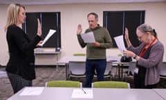 Three white people stand in a semicircle in a conference room with shades drawn, holding pieces of paper, holding their right hands up, and all appearing to speak at the same time.