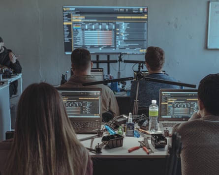 Women and men in a classroom facing a screen sit at desks on which open laptops show technical specifications. Next to the laptops are tools and drone components