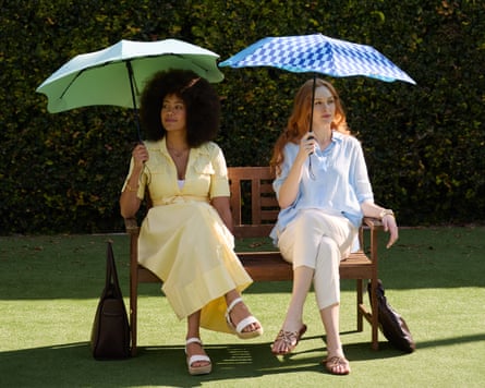 Two women in summer clothes sit on a bent on grass with stylish umbrellas overhead