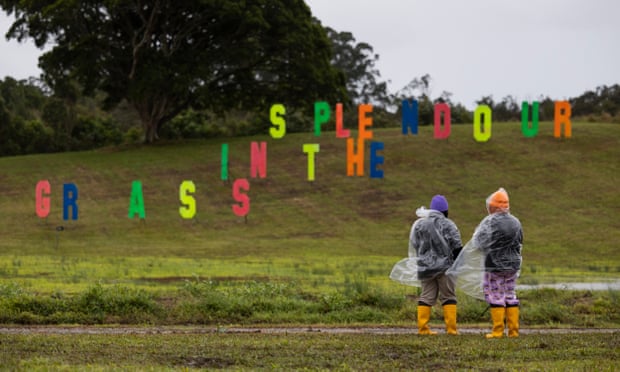 Two people in gumboots and rain ponchos stand in a field looking out to the rainbow-coloured Splendour in the Grass sign on a grass hill