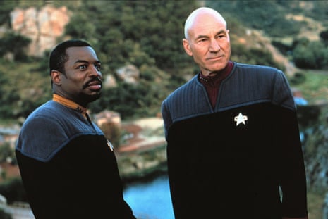 Geordi La Forge and Captain Picard stand outdoors in Starfleet uniforms, one glancing sideways