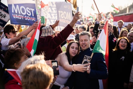 A young woman holds up a phone in front of herself and Magyar as they stand in a crowd at a rally. People are cheering and waving flags around them.