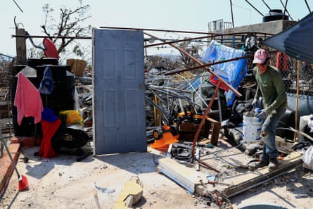 A man stands on what appears to be a concrete pillar in front of wreckage from a home, including a single still-standing door