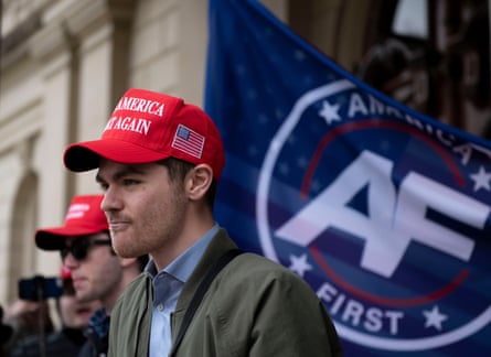A young man wearing a red Maga hat in front of an “America First” flag