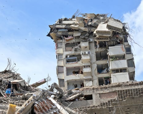 A heavily damaged multi-story apartment building with exposed interiors stands amid rubble and collapsed concrete structures