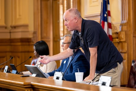 Man in polo shirt and khakis on dias stands, with two people seated beside him.