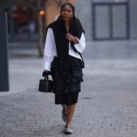 Kenya Hunt walks in the street wearing long transparent earrings, a black wool pullover around the neck, a white skirt, a black knee-length skirt and and rhinestones-covered black leather flats. She is carrying a black leather Chanel case