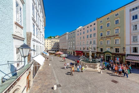 people walk in a wide square between tall pastel-coloured buildings. There is a fountain, and market stalls in the background.