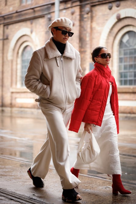 A man in a beige cap, jacket and white pants, walking alongside a woman in a white dress and a red puffer jacket draped over her shoulders.