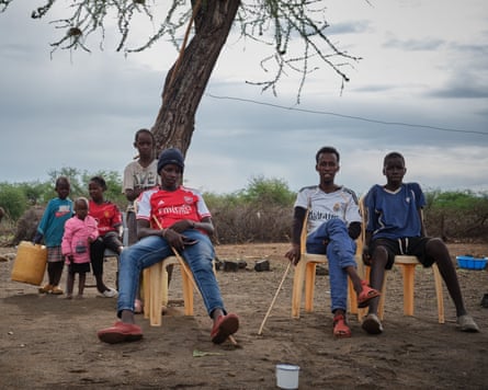 A group of children sit on plastic chairs under a bare tree.