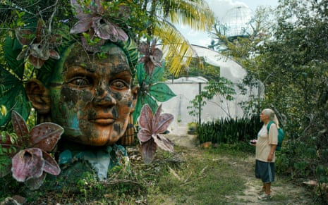 A woman stands looking at a huge sculpture of a head covered in plant growth.