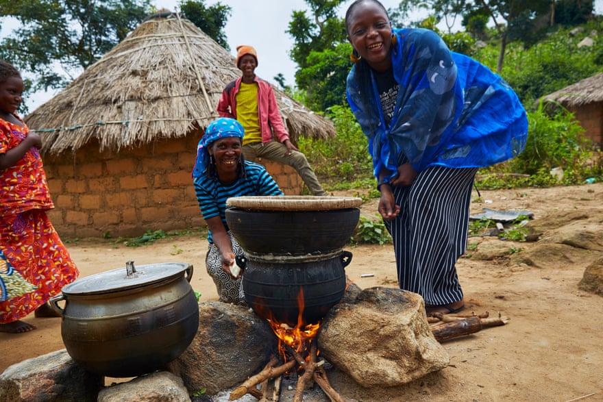 A young woman with an older woman at an open fire with a large cooking pot in front of a traditional African hut