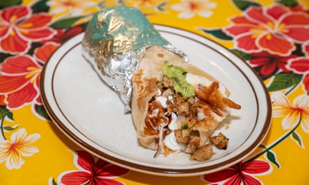 A chicken burrito with sour cream and guacamole sits on a white plate, half wrapped in foil, atop a yellow tablecloth with red and white flowers. 